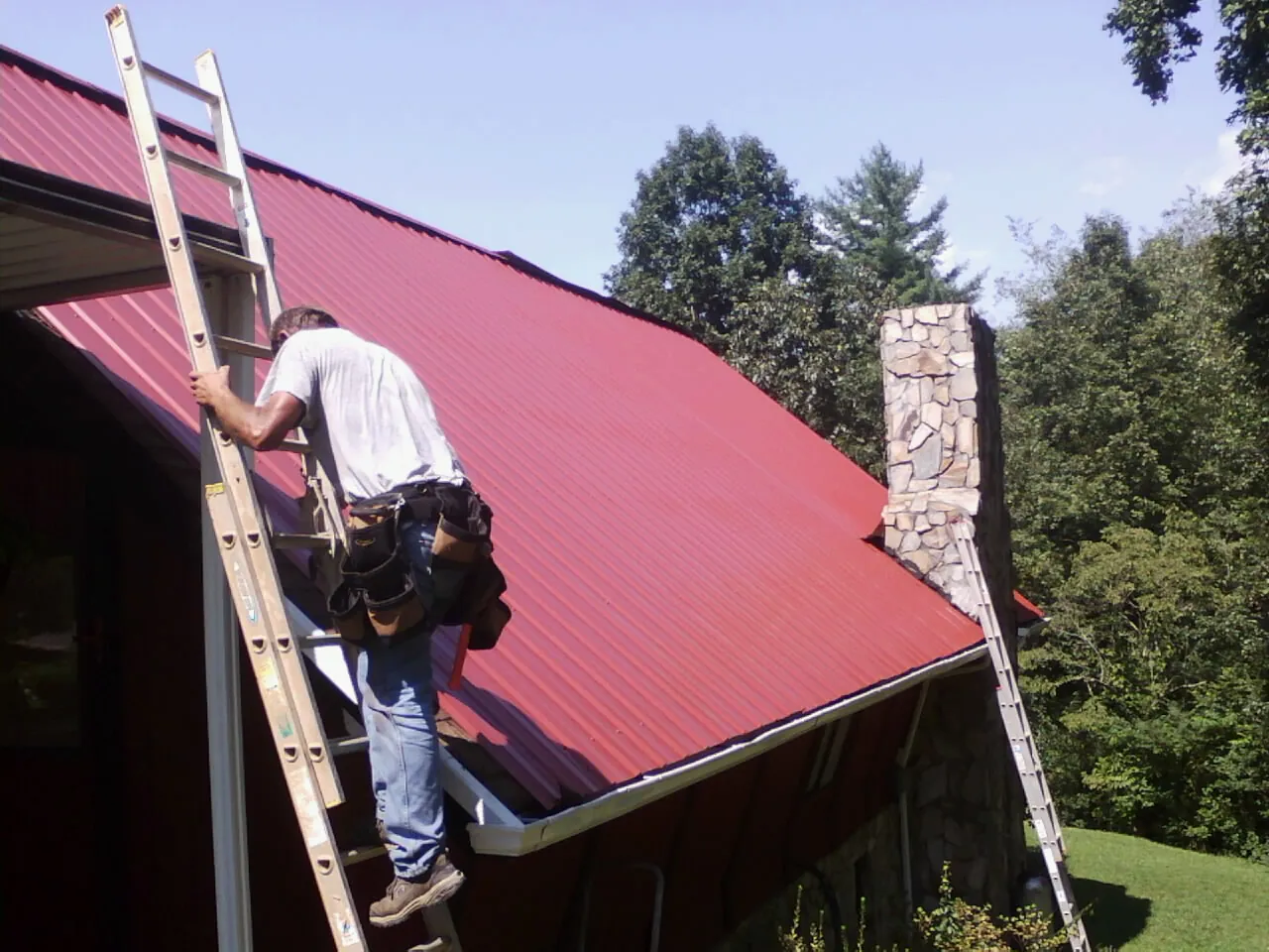 Roofer on a ladder beside a red metal roof and stone chimney — Soffit & Fascia Repair by Roofing Brothers in Terreton